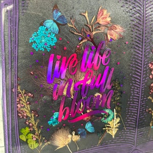 Op de afbeelding: Een paars boekvormig kunstharswerk met gedroogde bloemen en de tekst "live life in full bloom" in een verloop van roze en paars.