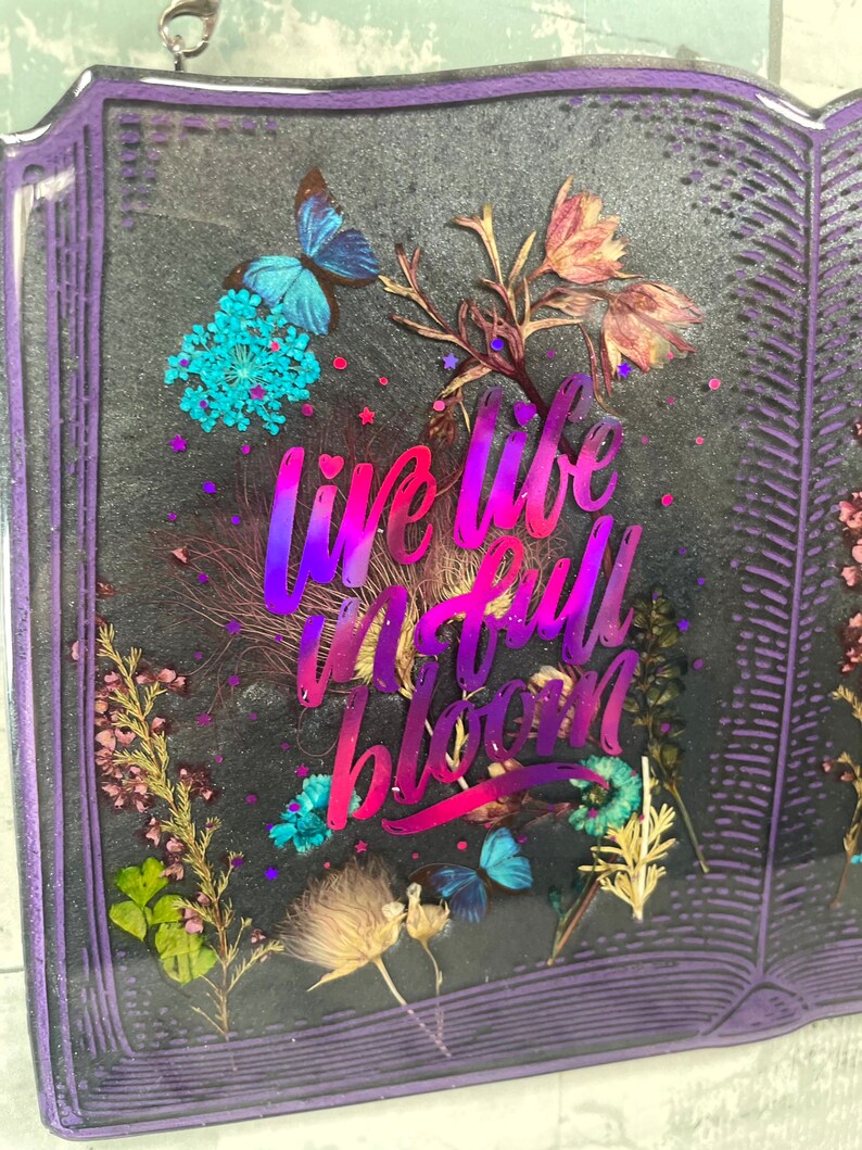 Op de afbeelding: Een paars boekvormig kunstharswerk met gedroogde bloemen en de tekst "live life in full bloom" in een verloop van roze en paars.