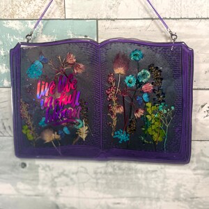 Op de afbeelding: Een paarse, boekvormige wanddecoratie van hars met gedroogde bloemen en de tekst "Live life in full bloom".