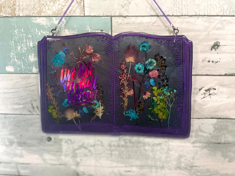 Op de afbeelding: Een paarse, boekvormige wanddecoratie van hars met gedroogde bloemen en de tekst "Live life in full bloom".