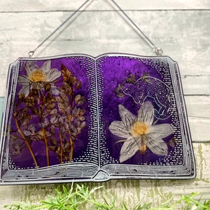 May include: A purple book-shaped resin ornament with pressed flowers and a white silhouette of a chameleon. The book is hanging from a silver chain.