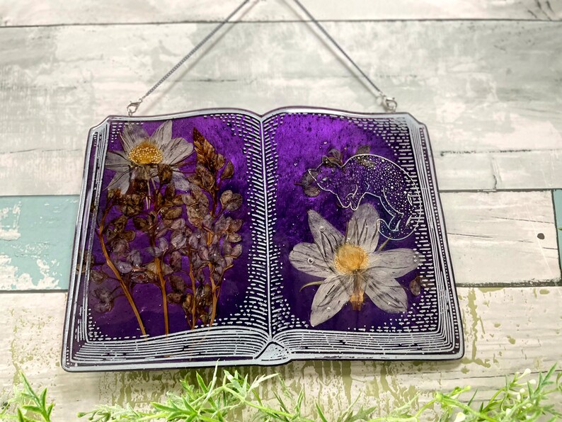 May include: A purple book-shaped resin ornament with pressed flowers and a white silhouette of a chameleon. The book is hanging from a silver chain.