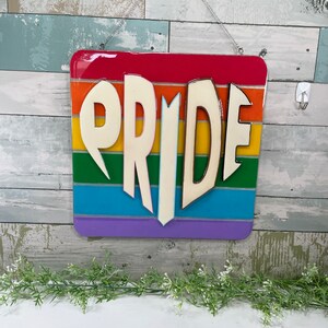May include: A rainbow-colored wooden sign with the word "PRIDE" in white letters. The letters are raised and have a 3D effect.
