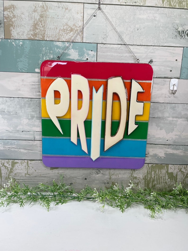 May include: A rainbow-colored wooden sign with the word "PRIDE" in white letters. The letters are raised and have a 3D effect.