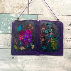 Op de afbeelding: Paarse, boekvormige wanddecoratie van hars met gedroogde bloemen en de tekst "Live life in full bloom".