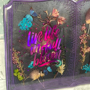 Op de afbeelding: Een paars boekvormig kunstharswerk met gedroogde bloemen en de tekst "live life in full bloom" in een holografisch roze en paarse lettertype.