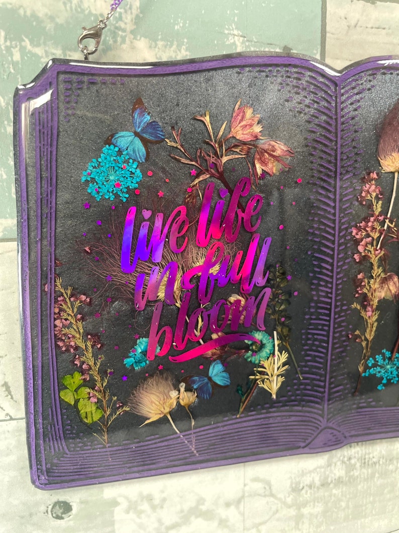 Op de afbeelding: Een paars boekvormig kunstharswerk met gedroogde bloemen en de tekst "live life in full bloom" in een holografisch roze en paarse lettertype.