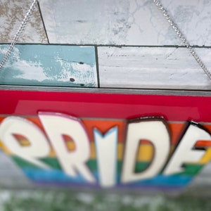 May include: A rainbow-colored wooden sign with the word "PRIDE" in white raised letters. The sign is hanging from a silver chain.