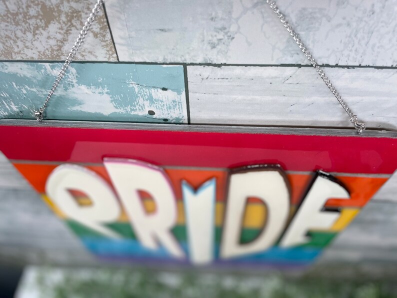May include: A rainbow-colored wooden sign with the word "PRIDE" in white raised letters. The sign is hanging from a silver chain.