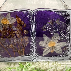 May include: A purple book-shaped resin ornament with a white border and a pressed flower design. The book is open to reveal a white flower and a black cat with white spots.