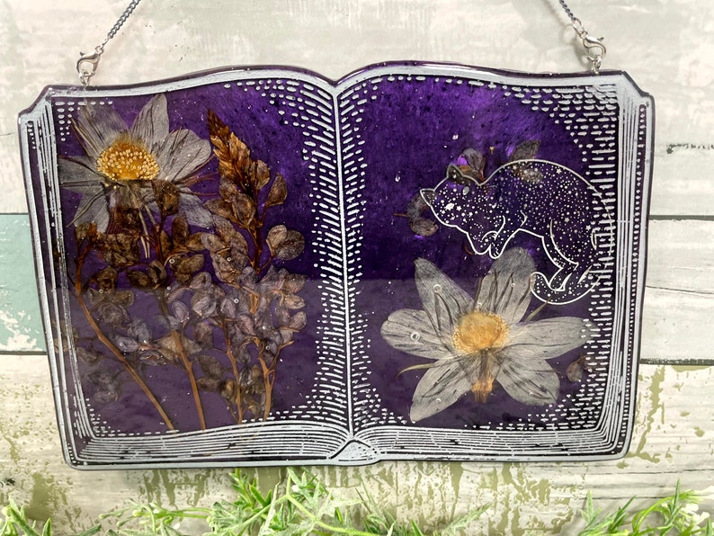 May include: A purple book-shaped resin ornament with a white border and a pressed flower design. The book is open to reveal a white flower and a black cat with white spots.