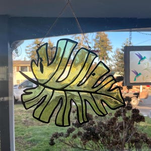 May include: A stained glass window hanging with a chain. The window is shaped like a large green leaf with black outlines.
