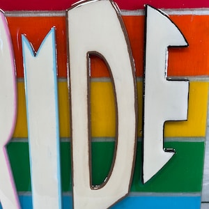 May include: Close-up of the letters "RI" from the word "PRIDE" painted in white with brown outlines on a rainbow striped background.