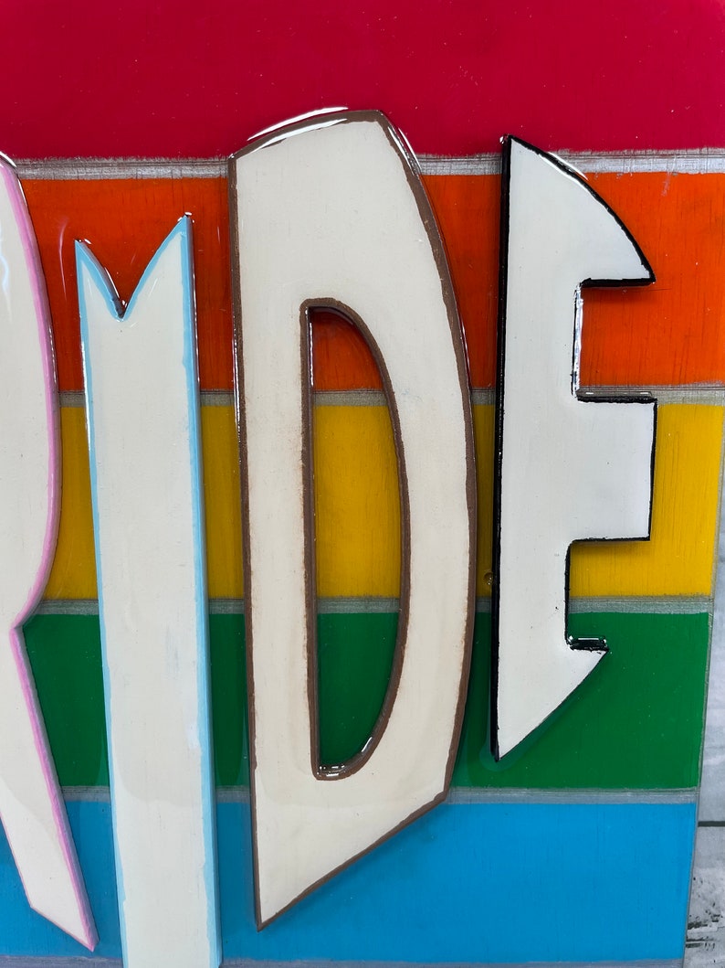 May include: Close-up of the letters "RI" from the word "PRIDE" painted in white with brown outlines on a rainbow striped background.