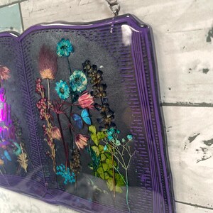 Op de afbeelding: Een paarse, boekvormige wanddecoratie van hars met een ontwerp van geperste bloemen. De bloemen zijn een mix van blauw, roze en groen. Het boek heeft een zilveren ketting die aan de bovenkant is bevestigd om op te hangen.