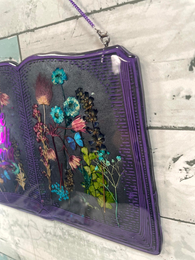 Op de afbeelding: Een paarse, boekvormige wanddecoratie van hars met een ontwerp van geperste bloemen. De bloemen zijn een mix van blauw, roze en groen. Het boek heeft een zilveren ketting die aan de bovenkant is bevestigd om op te hangen.
