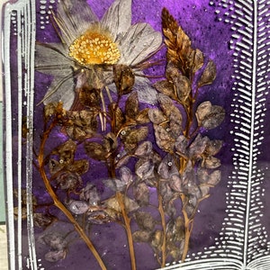 May include: A purple book with a white border and a pressed flower design. The flowers are white, brown, and purple.