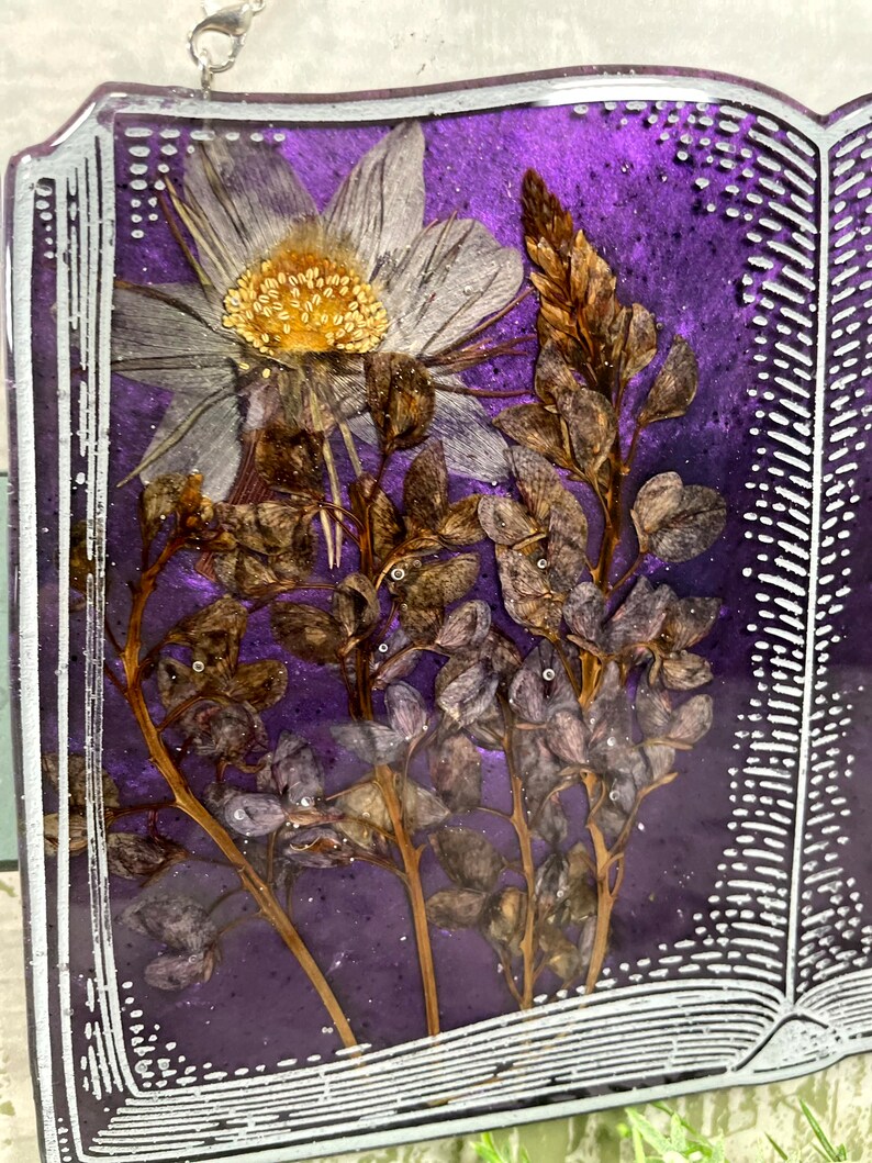 May include: A purple book with a white border and a pressed flower design. The flowers are white, brown, and purple.