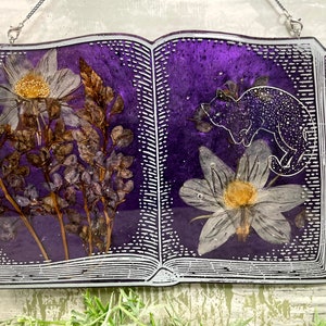 May include: A purple book with a white outline and a pressed flower design. The book is open to reveal a white flower and a black cat silhouette.