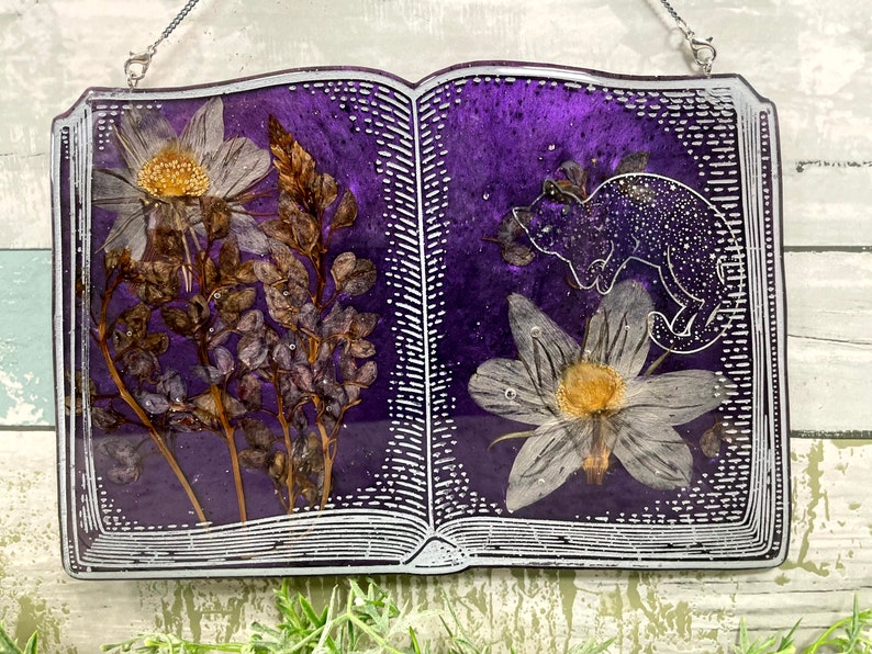 May include: A purple book with a white outline and a pressed flower design. The book is open to reveal a white flower and a black cat silhouette.