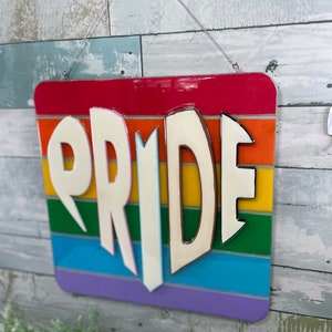 May include: A wooden sign with the word "PRIDE" in white letters on a rainbow background. The sign is hanging from a chain.