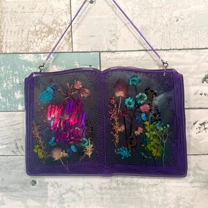 Op de afbeelding: Een paarse, boekvormige wanddecoratie van hars met gedroogde bloemen en de tekst "Live life in full bloom" in een regenboogkleurige gradi&euml;nt.