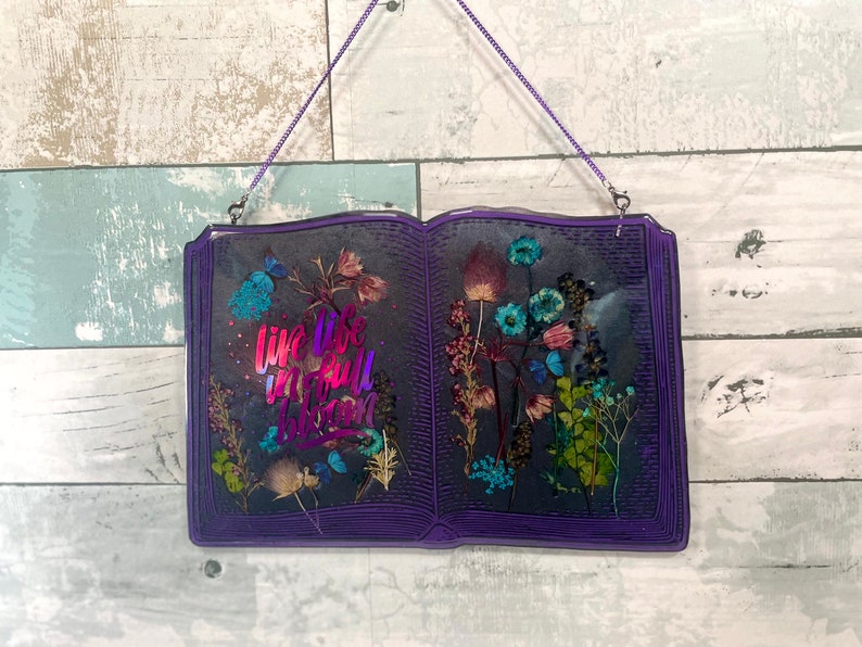 Op de afbeelding: Een paarse, boekvormige wanddecoratie van hars met gedroogde bloemen en de tekst "Live life in full bloom" in een regenboogkleurige gradi&euml;nt.