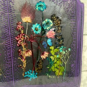 Op de afbeelding: Een paars boekvormig kunstharswerk met geperste bloemen en vlinders erin. De bloemen zijn in tinten roze, blauw, groen en bruin. De vlinders zijn blauw en wit.