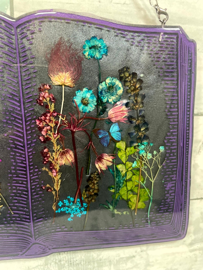 Op de afbeelding: Een paars boekvormig kunstharswerk met geperste bloemen en vlinders erin. De bloemen zijn in tinten roze, blauw, groen en bruin. De vlinders zijn blauw en wit.