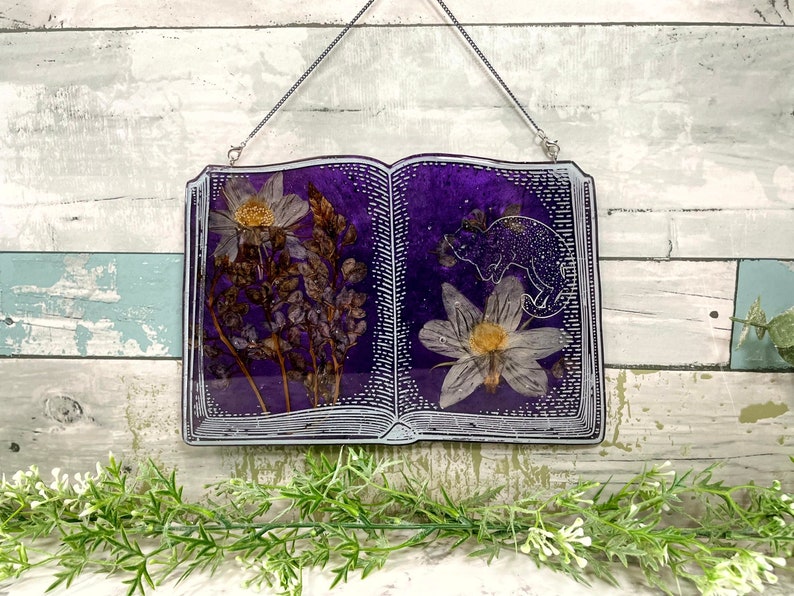 May include: A purple book-shaped resin wall hanging with a white frame and a pressed flower design. The book is open to reveal a white flower and a small, white, glittery animal silhouette.