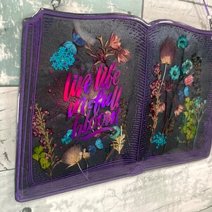 Op de afbeelding: Paarse boekvormige kunstharskunst met gedroogde bloemen en de tekst "Live life in full bloom" in een regenboogkleurige gradi&euml;nt.