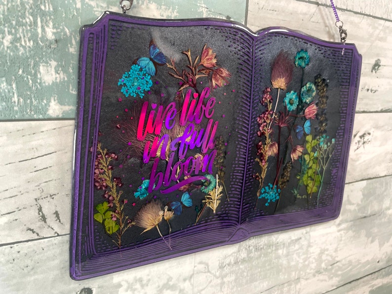 Op de afbeelding: Paarse boekvormige kunstharskunst met gedroogde bloemen en de tekst "Live life in full bloom" in een regenboogkleurige gradi&euml;nt.