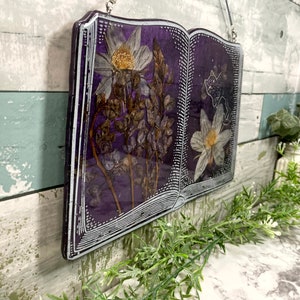 May include: A purple book-shaped resin art piece with pressed flowers and a white silhouette of a cat. The flowers are white and brown and the book is framed with a white border.
