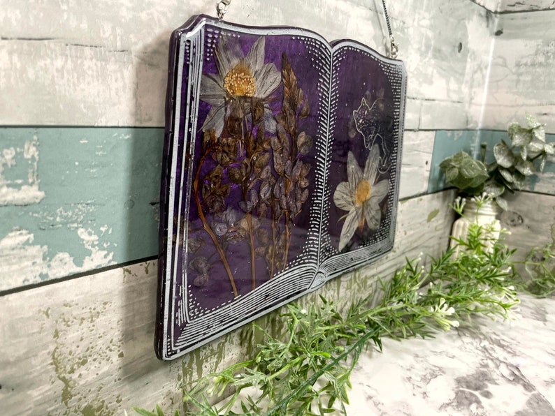 May include: A purple book-shaped resin art piece with pressed flowers and a white silhouette of a cat. The flowers are white and brown and the book is framed with a white border.
