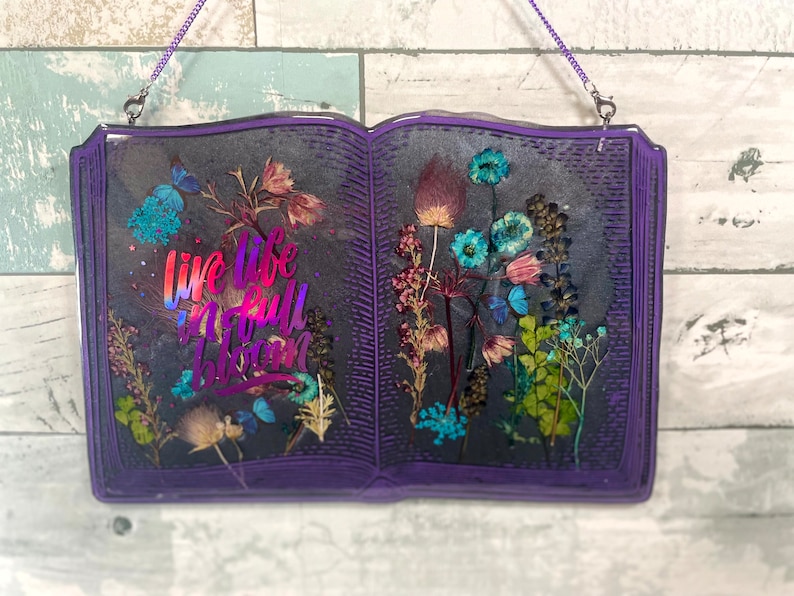 Op de afbeelding: Een paarse, boekvormige wanddecoratie van hars met gedroogde bloemen en de tekst "Live life in full bloom" in iriserende letters.