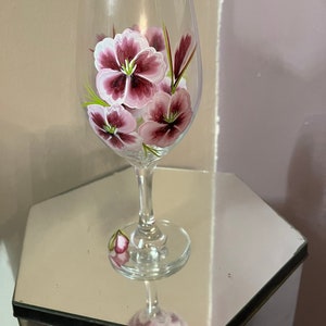 May include: A clear glass wine glass with a hand-painted design of pink and purple flowers. The flowers have white centers and green stems.