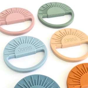May include: A collection of colorful, circular baby teethers in shades of pink, green, blue, and orange. Each teether features a sunburst design and a handle. Some teethers have names like "Jonas" and "Emma" inscribed.