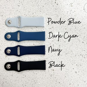 Pu&ograve; includere: Quattro cinturini per orologi in azzurro polvere, ciano scuro, blu navy e nero. Ogni cinturino ha un perno color argento e i nomi dei colori sono scritti in corsivo. I cinturini sono disposti su una superficie bianca maculata.