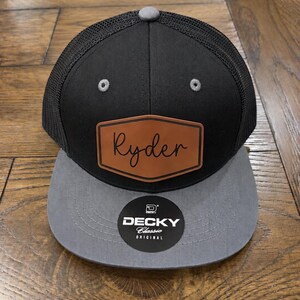 May include: A black and grey trucker hat featuring a black mesh back and a solid black front panel. A brown leather-like patch is centered on the front with the name Ryder in black script. A round black sticker reads DECKY Classic ORIGINAL.