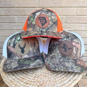 May include: Three trucker caps featuring a camouflage pattern. The top cap has bright orange mesh and a circular leather patch with a fish. The bottom left cap has white mesh and a deer head patch. The bottom right cap has white mesh and a duck patch.