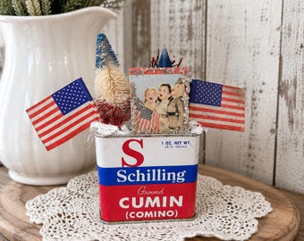 Può includere: Una scatola vintage di cumino macinato Schilling con marchio rosso, bianco e blu, etichettata 1 OZ. NET WT. 28.35 GRAMS. È decorata con due bandiere americane in miniatura, un piccolo albero a spazzola con setole rosse, bianche e blu e una piccola immagine.