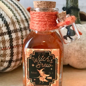 Pu&ograve; includere: Una piccola bottiglia di vetro con un tappo di sughero e un'etichetta che recita "Witch's Brew Potion No. 2" con una strega che vola su una scopa. La bottiglia &egrave; legata con uno spago arancione e ha una bottiglia di vetro pi&ugrave; piccola con un corvo nero all'interno che pende dal collo.
