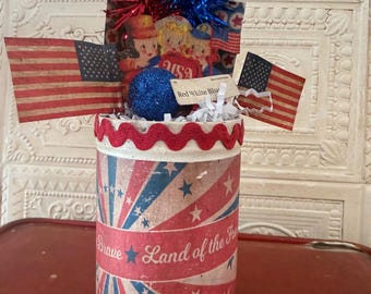 Può includere: Un contenitore cilindrico decorato con motivi a stella rossi, bianchi e blu e il testo Brave Land of the Free. Presenta due bandiere americane, festoni rossi e blu, una sfera blu glitterata e un'etichetta con scritto Red White Blue.