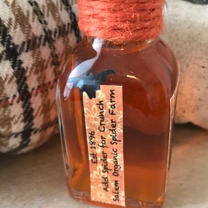 Pu&ograve; includere: Una piccola bottiglia di vetro con un liquido marrone al suo interno. La bottiglia &egrave; legata con uno spago e ha un'etichetta che recita "Est 1896 Add Spider for Crunch Salem Organic Spider Farm".