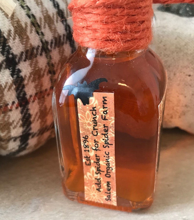 Pu&ograve; includere: Una piccola bottiglia di vetro con un liquido marrone al suo interno. La bottiglia &egrave; legata con uno spago e ha un'etichetta che recita "Est 1896 Add Spider for Crunch Salem Organic Spider Farm".