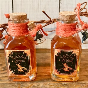 Pu&ograve; includere: Due piccole bottiglie di vetro con tappi di sughero e spago arancione legato attorno al collo. Le bottiglie sono riempite con un liquido dorato e hanno etichette nere che recitano "Witch's Brew Potion No. 7".