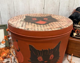 May include: A round, orange storage container with a lid featuring a black cat face design. The container has a checkered pattern on the lid and a pumpkin graphic. Decorative stars hang in the background.