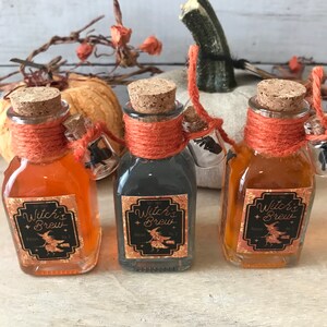 Pu&ograve; includere: Tre piccole bottiglie di vetro con tappi di sughero e spago arancione legato attorno al collo. Ogni bottiglia contiene un liquido di colore diverso e ha un'etichetta che recita "Witch's Brew" con un'illustrazione di una strega.