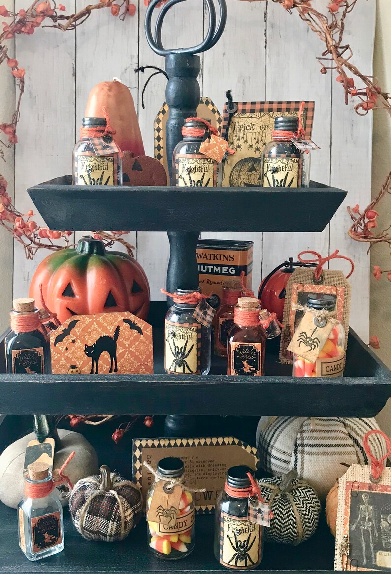 Pu&ograve; includere: Un vassoio a tre livelli nero decorato con oggetti a tema Halloween, tra cui piccole bottiglie di vetro con etichette che recitano "Frightful", "Witch's Brew" e "Candy Corn", insieme a zucche, un gatto nero e un cartello che dice "Trick or Treat".