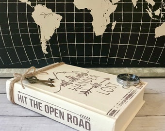Può includere: Una scatola di legno bianca a forma di libro con il testo "Hit the Open Road" e "Not All Who Wander Are Lost" sul davanti. La scatola ha una bussola e due chiavi attaccate.